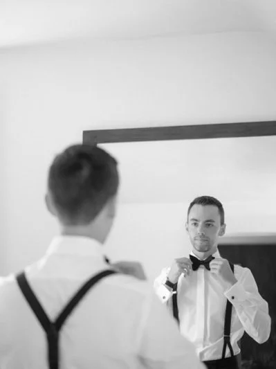 A man in a tuxedo adjusting his bow tie in front of a mirror, with another man’s reflection seen in the mirror.