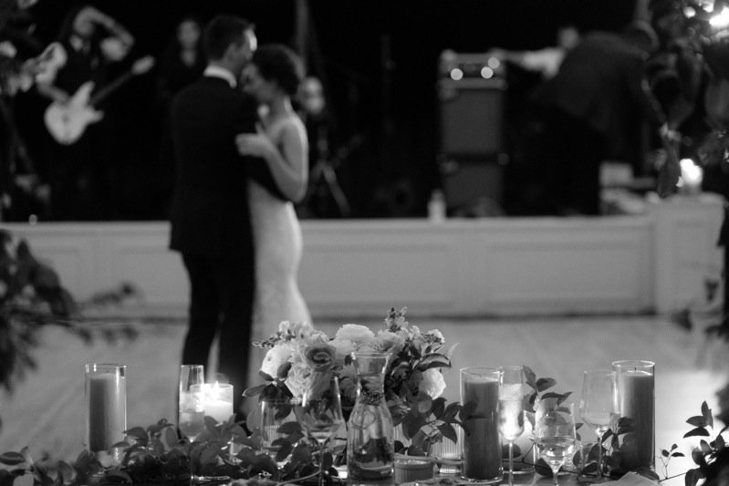 A black and white photo of a bride and groom dancing at their wedding reception, with a table of flowers and candles in the foreground.