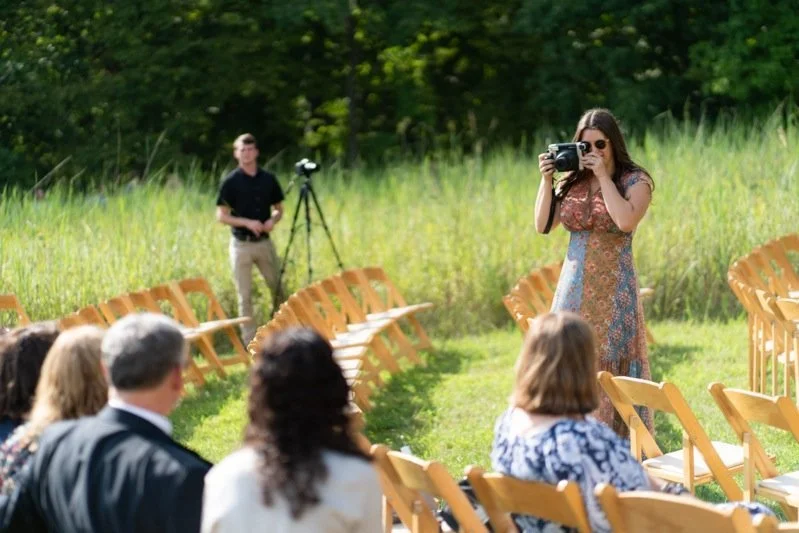 A woman with sunglasses taking photos at an outdoor event with several people seated in wooden chairs and a man near a camera tripod in the background.