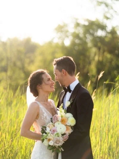 A bride and groom standing close together outdoors, with the bride holding a bouquet of flowers, during sunset.
