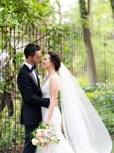 Bride and groom in wedding attire standing close together outdoors, with a black metal fence and green trees in the background.