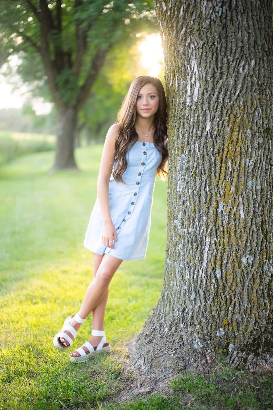 A young woman with long brown hair wearing a light blue dress and white sandals standing next to a large tree in a park during sunset.