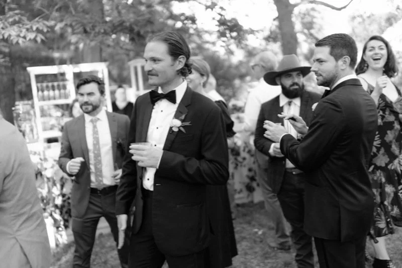 Group of people in formal attire at outdoor event, some holding drinks, trees in background