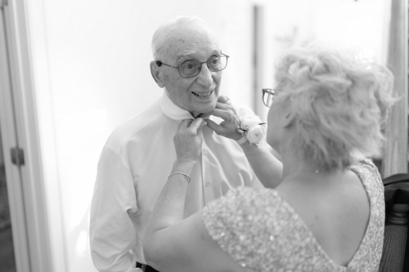 An elderly man with glasses smiling as a woman adjusts his bow tie.