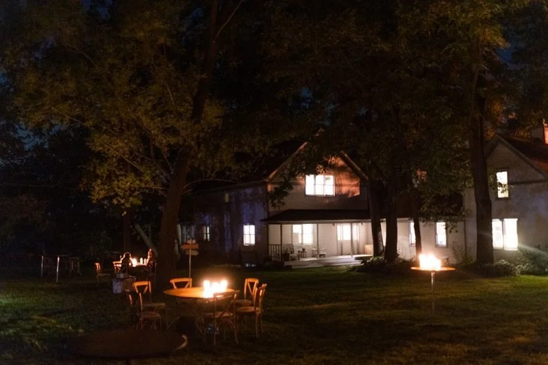 Nighttime backyard scene with lit fire pits and a house in the background.