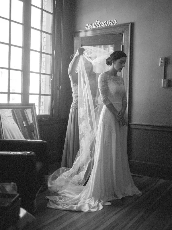 A bride in a wedding dress standing with her hands clasped in front of her inside a room, while a woman with a veil adjusts her hair or dress behind her. There is a window, a mirror, and a sign that says 'restrooms' above a door in the background.