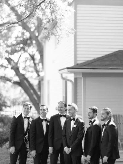 Six men dressed in black tuxedos and bow ties standing outdoors in front of a house, smiling.