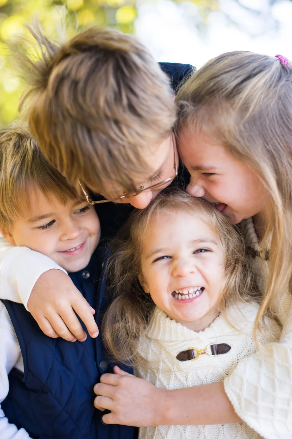 A group hug of a woman and three children outdoors, all laughing and smiling.