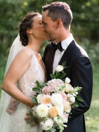 Bride and groom sharing a kiss on their wedding day, with the bride holding a bouquet of flowers and both dressed in formal wedding attire, outdoors in a green natural setting.