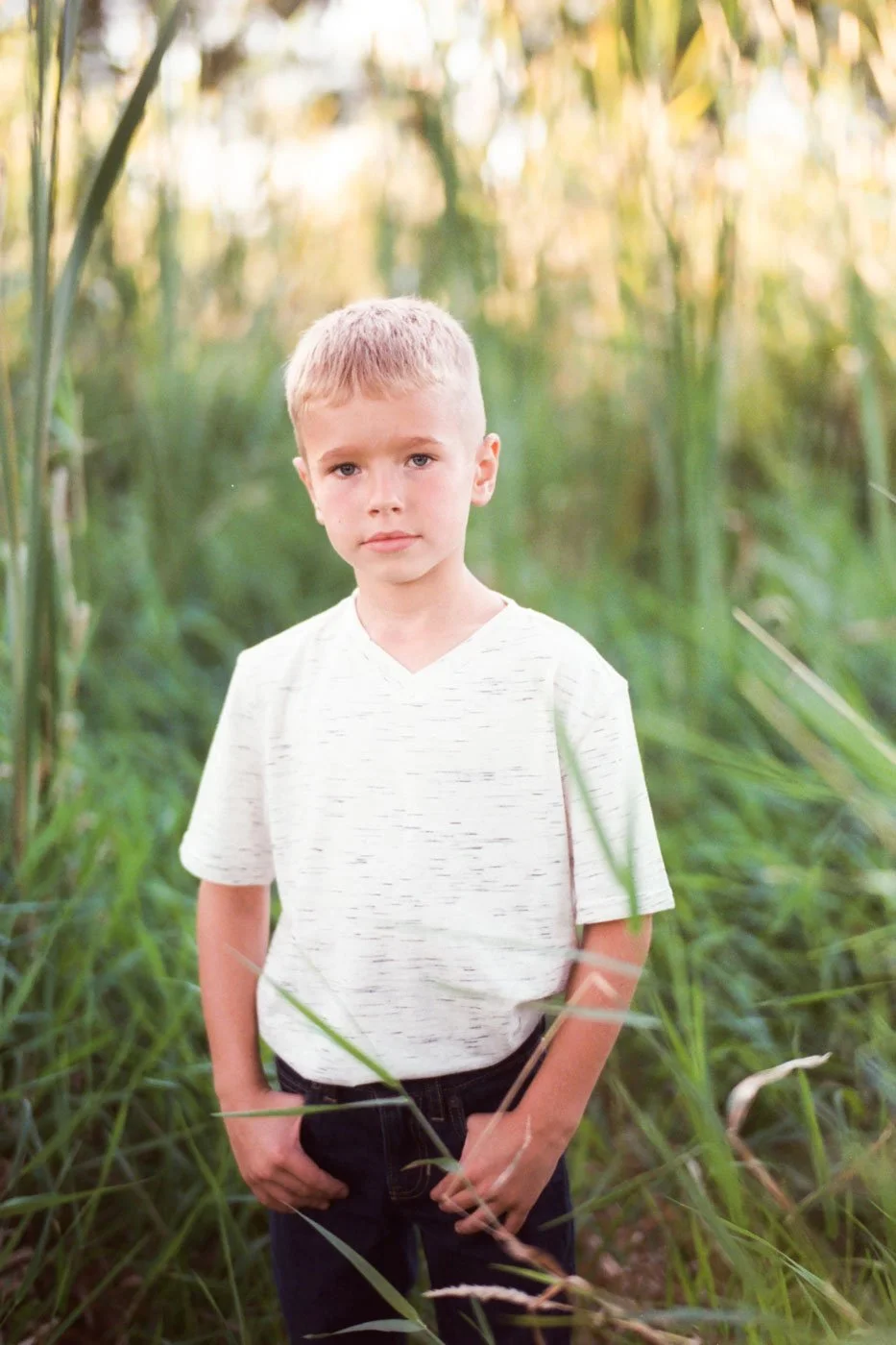 A young boy with blonde hair and blue eyes standing outdoors in a grassy area with tall green plants and trees in the background, wearing a white T-shirt and dark pants.