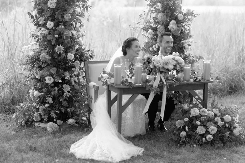 A newlywed couple sits at a wedding reception table outdoors, surrounded by floral arrangements, candles, and greenery.