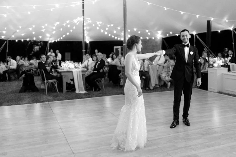 A bride and groom dance together at their wedding reception under string lights, with guests seated at tables watching.