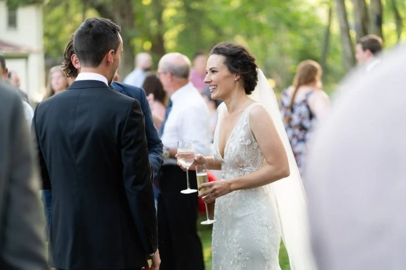 A bride in a white wedding dress and veil smiling and holding a glass of champagne, talking to a groom dressed in a black suit, at an outdoor wedding reception with others in the background.