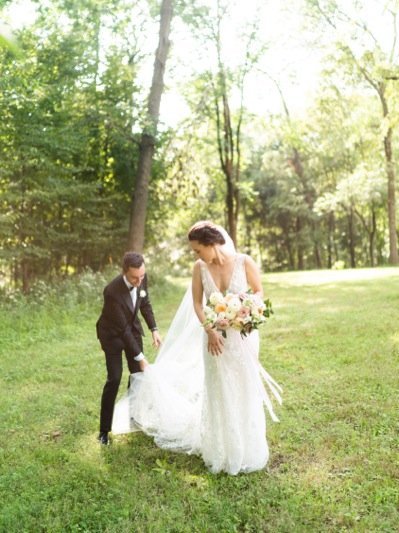 Bride and groom standing on grass in a park with trees, bride holding a bouquet, groom adjusting the bride's dress.