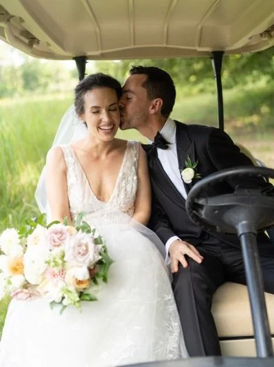 Bride and groom sitting in a golf cart, with the groom kissing the bride on her forehead, during a wedding photoshoot in a green outdoor setting.