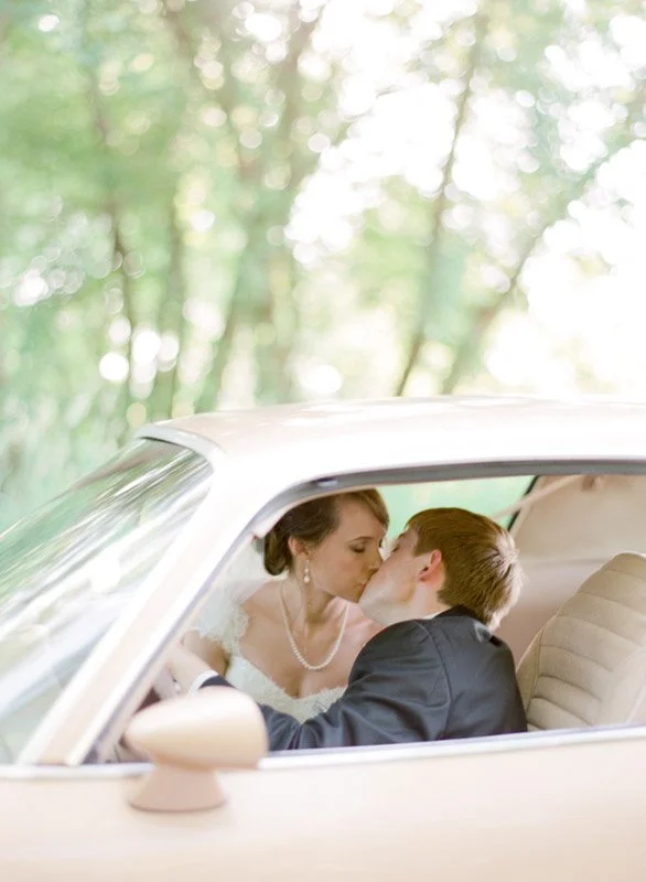 A bride and groom sharing a kiss inside a vintage white car with a blurred green forest background.