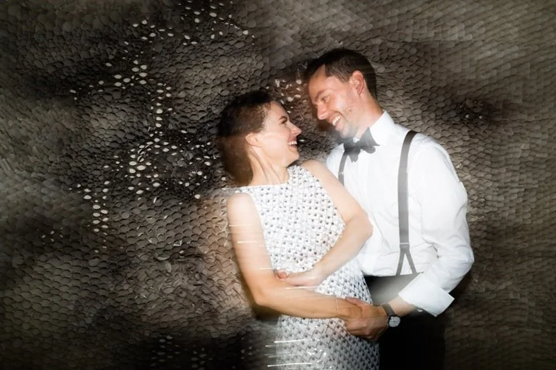 A smiling woman and man in formal attire sharing an intimate moment, holding hands, against a textured dark background.