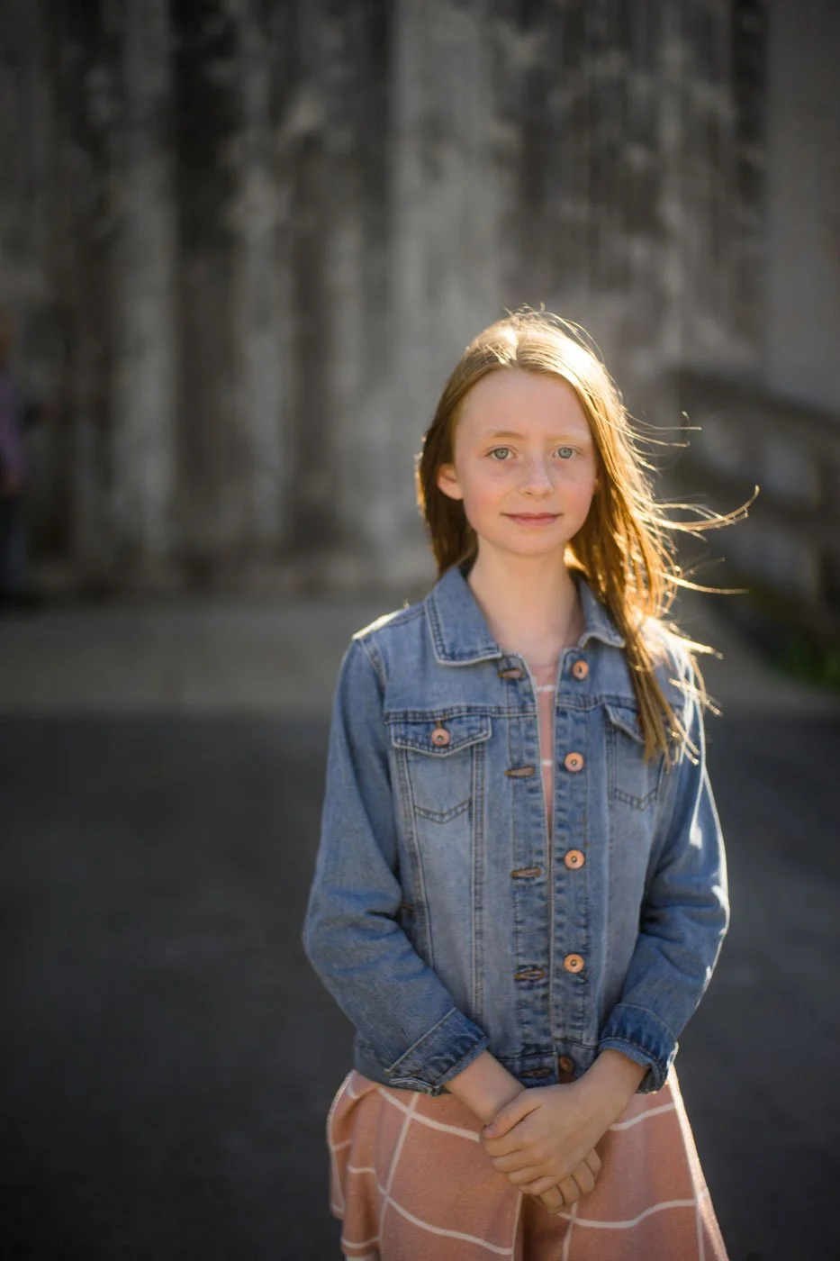 A young girl with red hair and blue eyes standing outdoors in front of a blurred stone wall, wearing a denim jacket and a peach-colored skirt, with sunlight shining on her hair.
