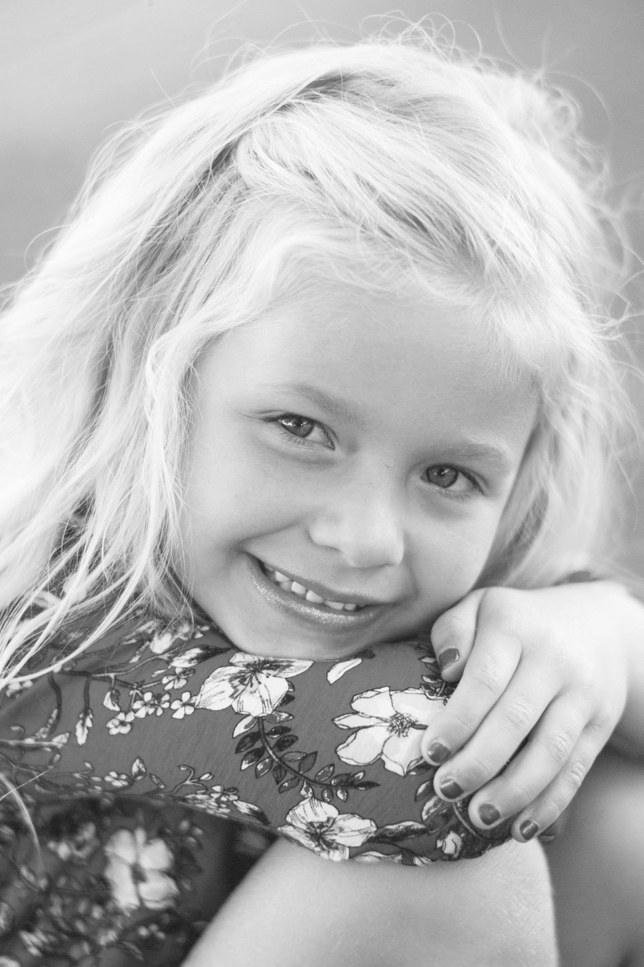 Black and white close-up photo of a young girl with blonde hair smiling and hugging someone, wearing a floral-patterned top.