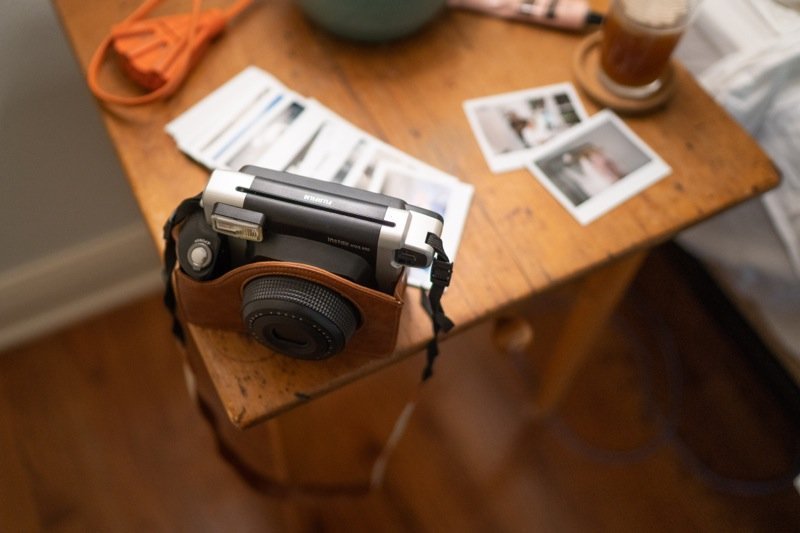 A digital camera with a brown cover hanging over the edge of a wooden table, with loose photographs, a glass of iced tea, and an orange bag on the table.