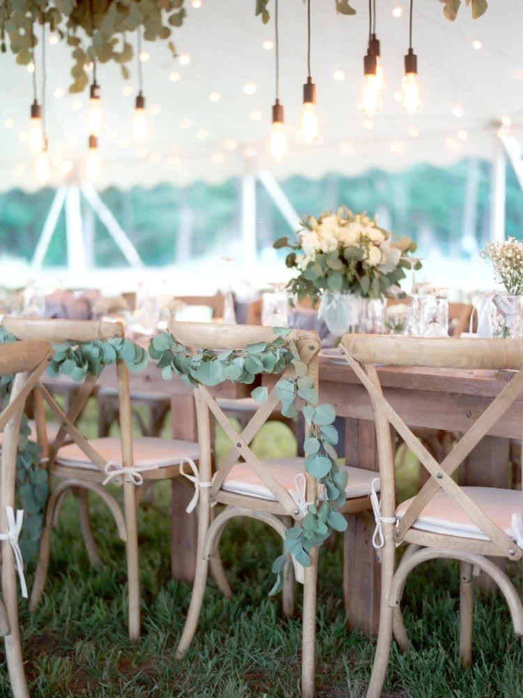 Outdoor wedding or event setup featuring wooden chairs with white cushions, decorated with eucalyptus garlands, around a rustic wooden table. The background shows a tent with hanging Edison bulbs and blurred trees outside.