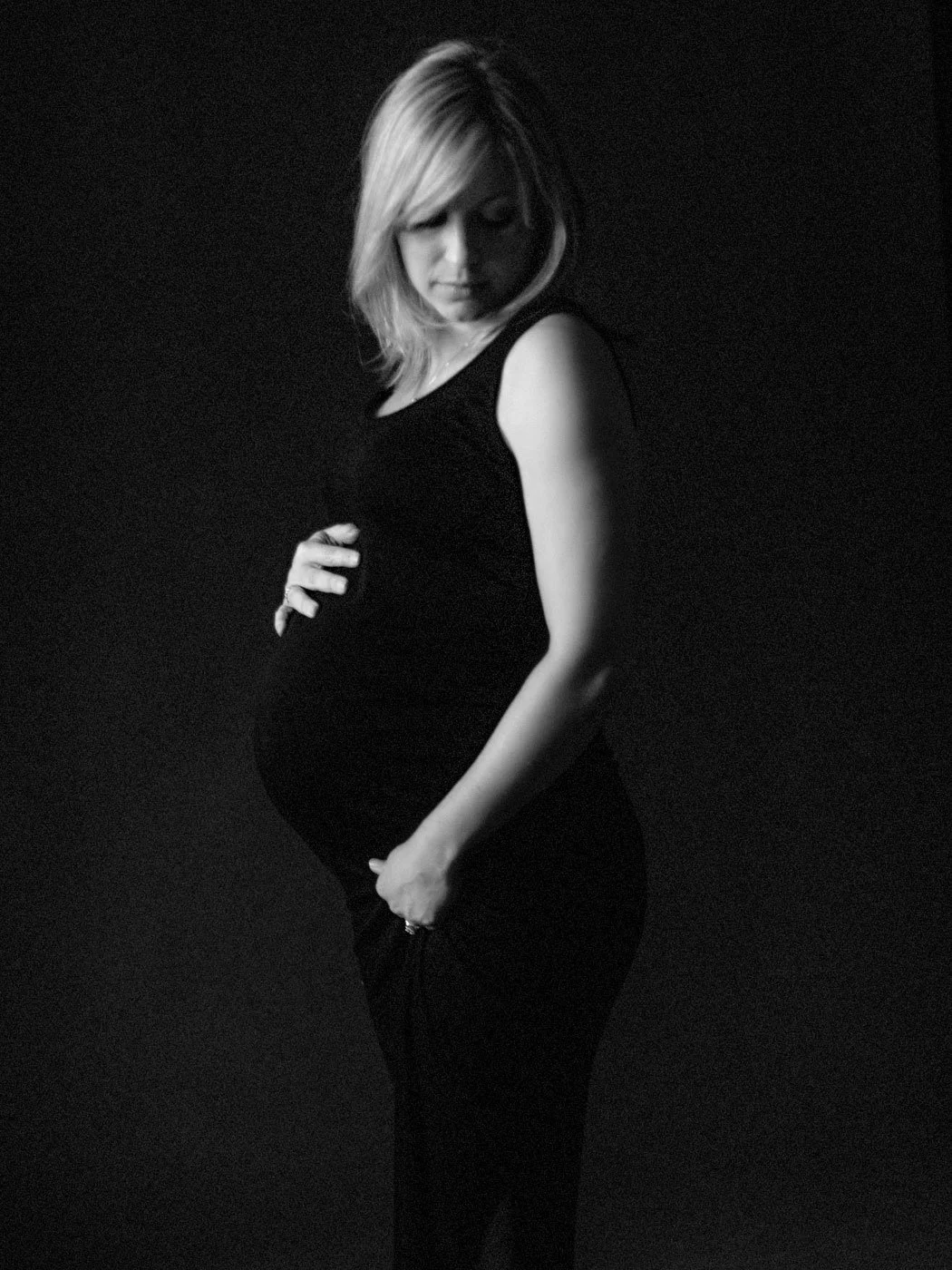 A pregnant woman in black clothing looking down, posed against a black background.