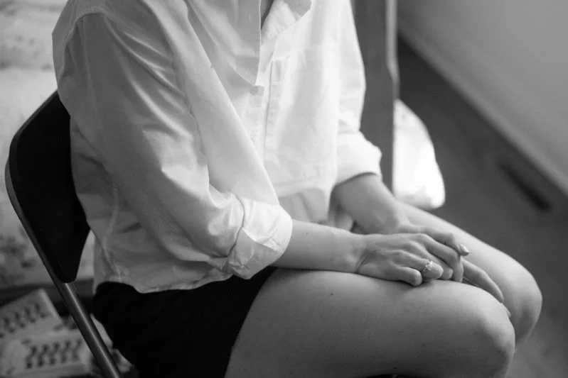 A person sitting on a chair with hands resting on their lap, wearing a white shirt and dark shorts, in a black-and-white photo.
