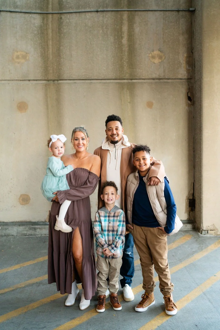 A diverse family of five standing together in front of a concrete wall, including a woman holding a young girl, a man, and three boys.