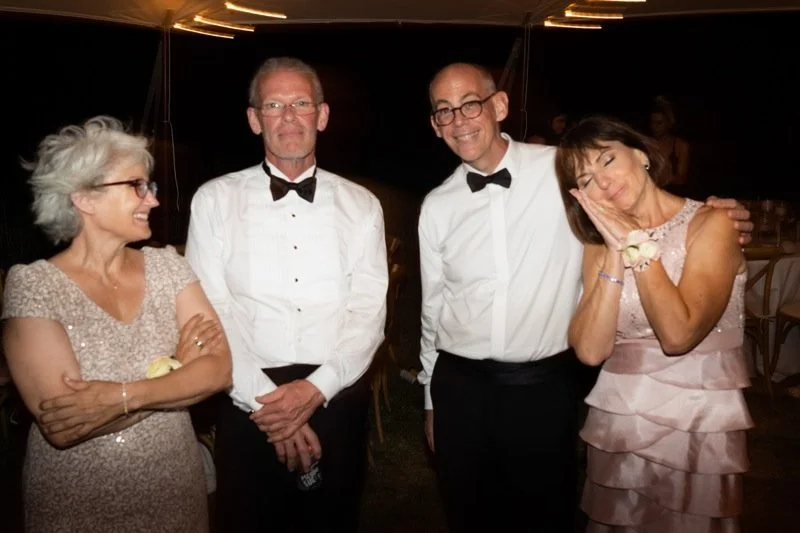Four people at a formal event, two women and two men, smiling and posing together, with a dark background and tables visible, suggesting a celebration or party.