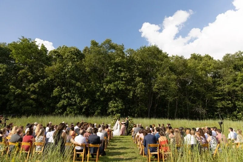 Outdoor wedding ceremony with a bride and groom exchanging vows in front of an audience, set against a backdrop of trees under a partly cloudy sky.