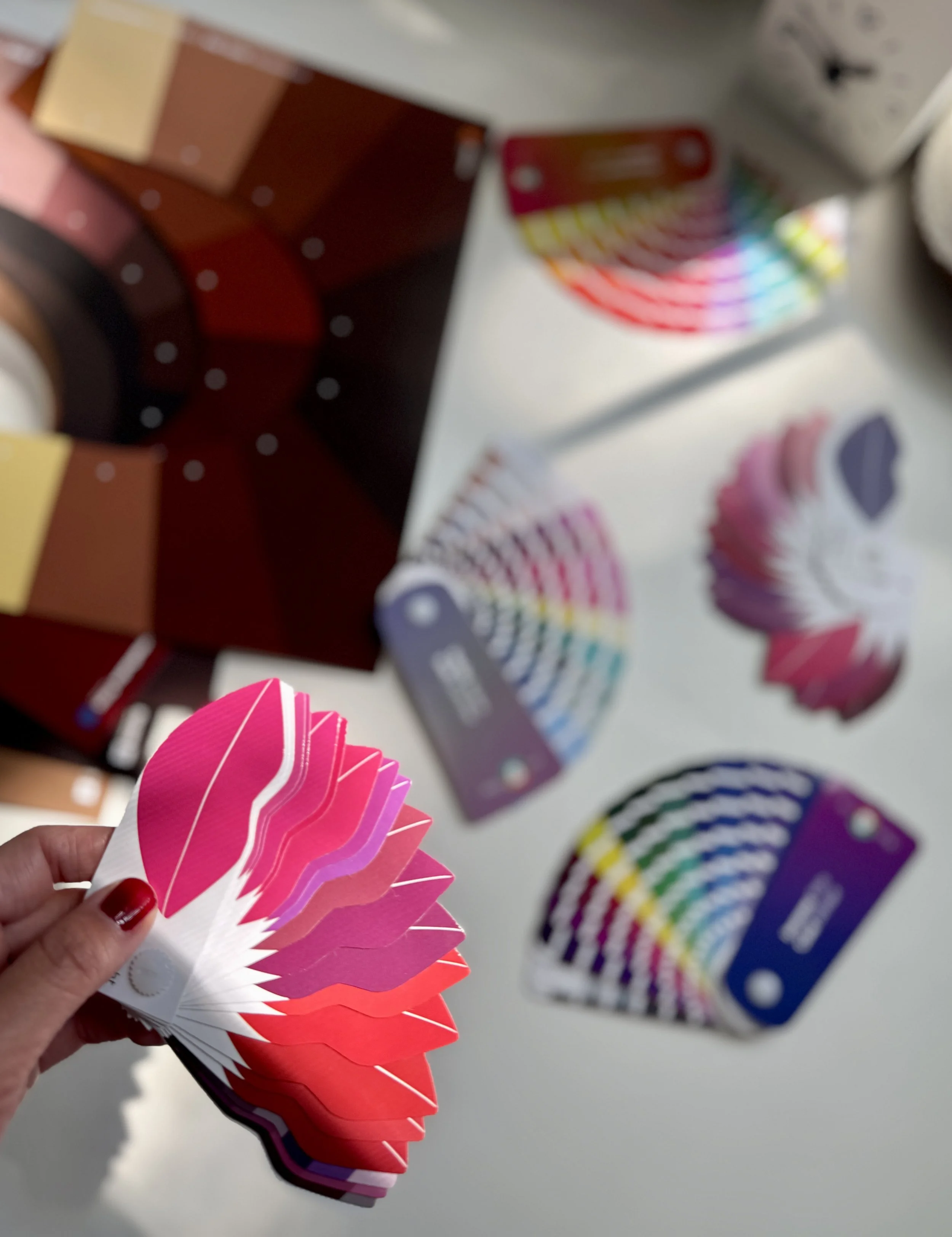 Color swatches on color fan deck held by a person with painted nails, along with other color fan decks and color sample cards on white surface.