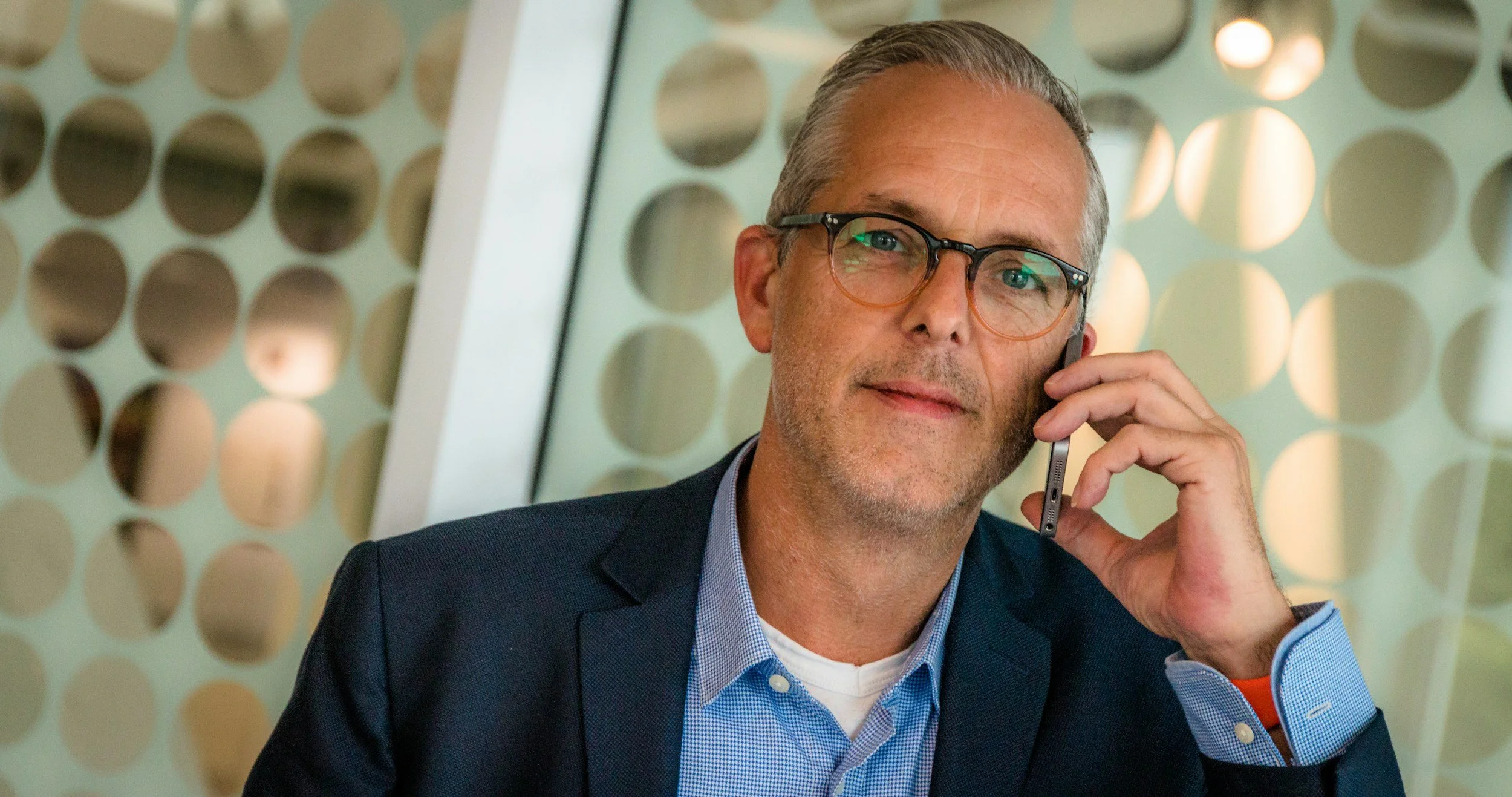 A middle-aged man with gray hair, glasses, wearing a navy blazer and light blue shirt, talking on a smartphone in an office with a modern, circular-patterned partition in the background.