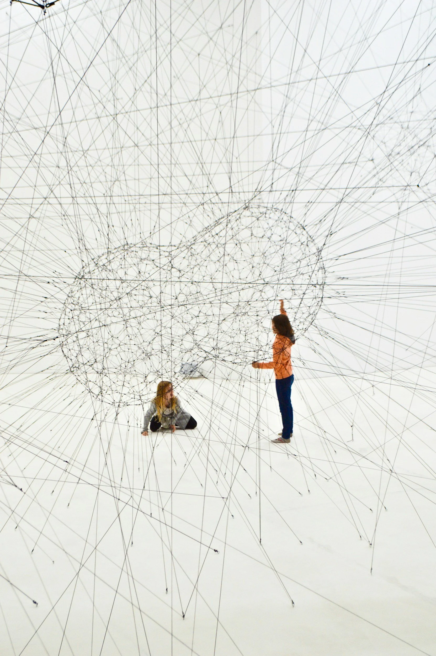 Two women interacting with an intricate web-like art installation made of thin black lines and small black nodes, set against a plain white background.
