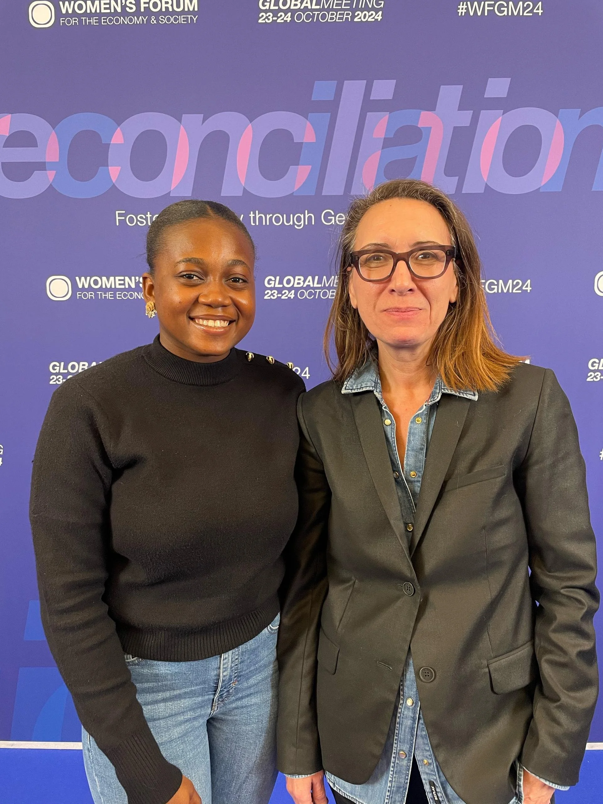 Two women standing together in front of a purple backdrop with text about the Women's Forum and global meeting, smiling at the camera.