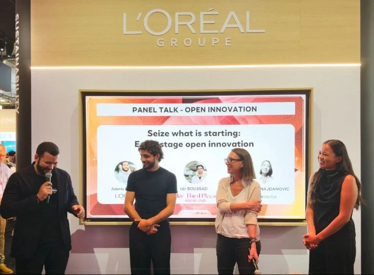 Four people on stage participating in a panel talk at an event, with a large screen behind them displaying the event's topic and speakers, and the L'Oréal Group logo at the top.