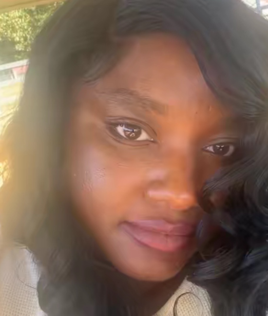 Close-up of a young woman with smooth skin and dark wavy hair, outdoors with green trees in the background.