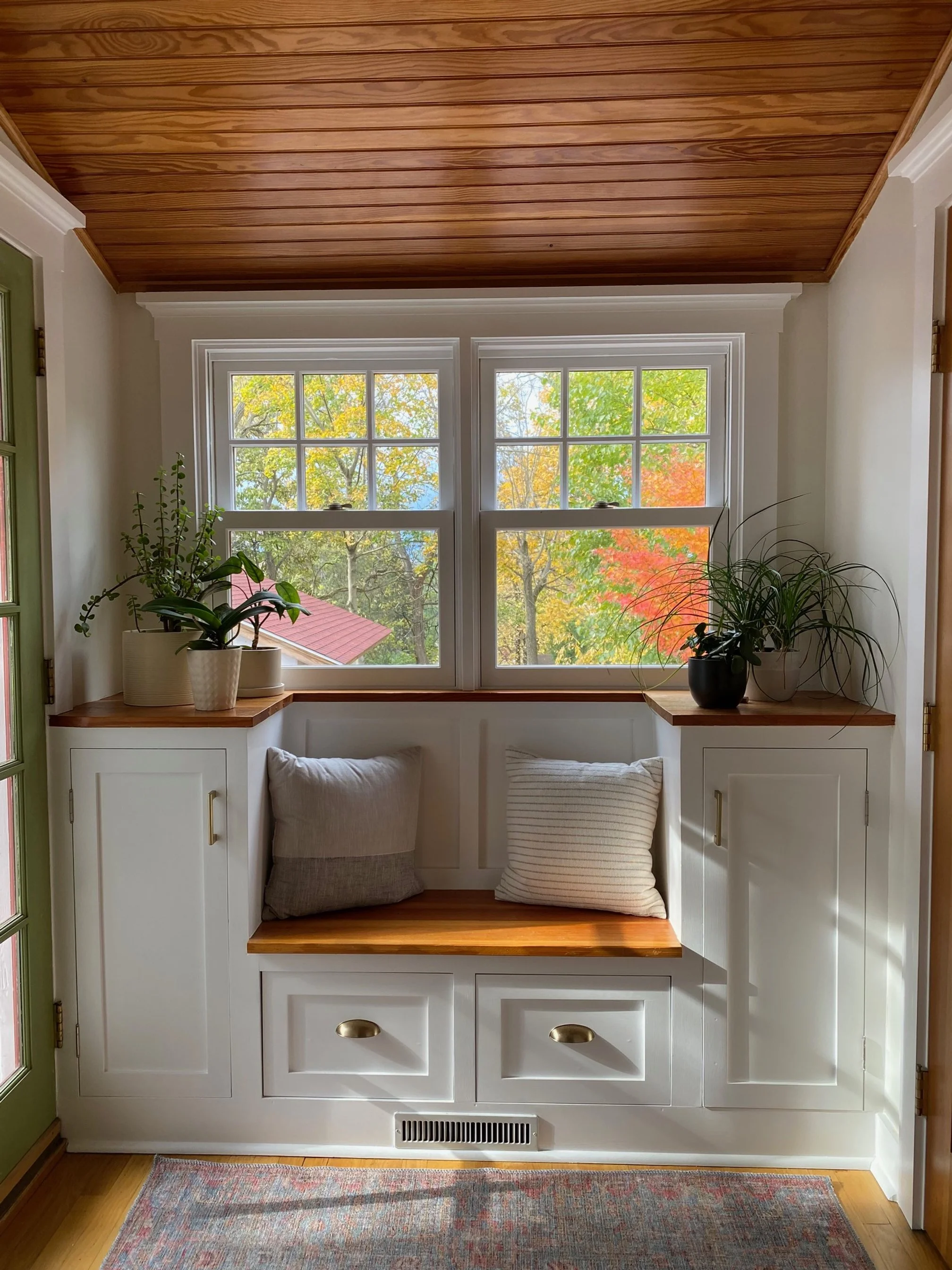 A cozy window nook with a built-in bench, white cabinetry with brass handles, pillows, potted plants on the window sill, and a view of colorful autumn trees outside.
