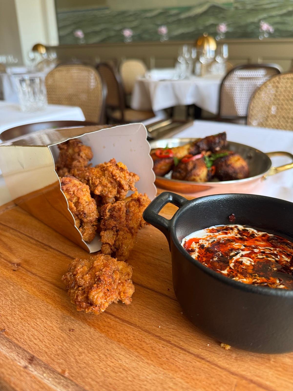 Fried chicken pieces with spicy dipping sauce, served on a wooden board in a restaurant dining area.