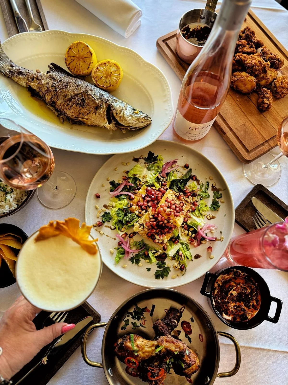 A table with various dishes including a whole grilled fish with lemon slices, a salad with pomegranate seeds, a small cast iron skillet with a cooked dish, and a bowl of chicken wings. There are also glasses of rosé wine and a cocktail with a pineapp