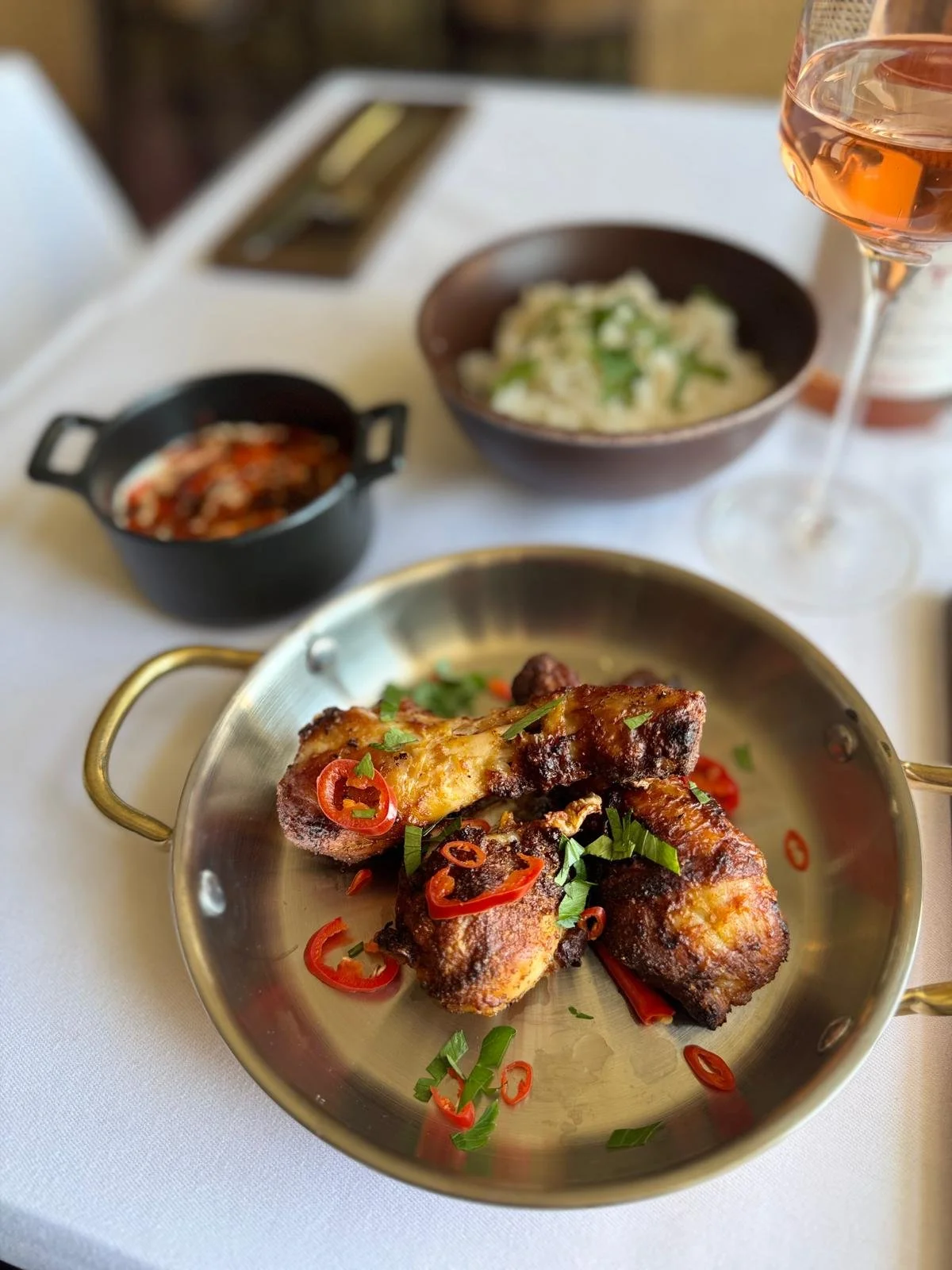 A plate of grilled chicken drumsticks garnished with sliced red chili peppers and chopped green onions, served on a small metallic tray. In the background, there is a bowl of mashed potatoes with green herbs, a bowl of chili, and a glass of rosé wine
