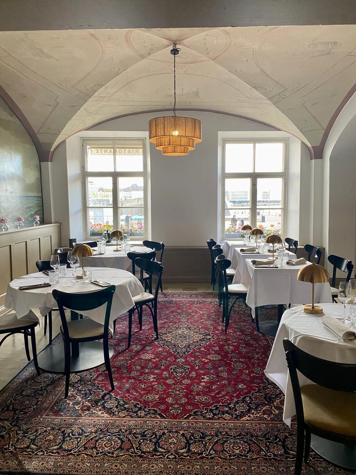 Elegant dining room with white tablecloth-covered tables, black chairs, gold table lamps, and large windows overlooking a waterfront scene, with decorative ceiling and a red patterned rug.