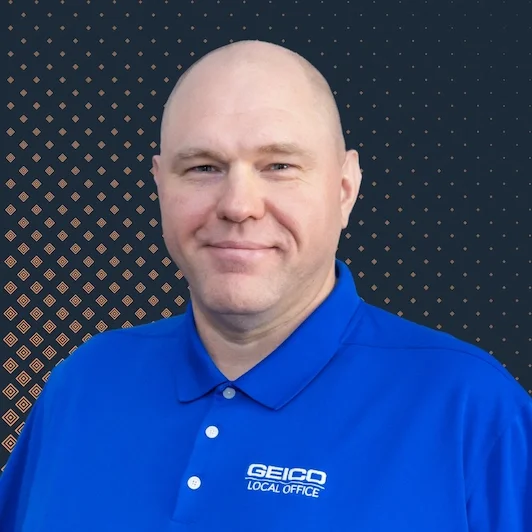 A man smiling, wearing a blue polo shirt with 'GEICO Local Office' logo, standing against a dark background with a pattern of small gold dots.