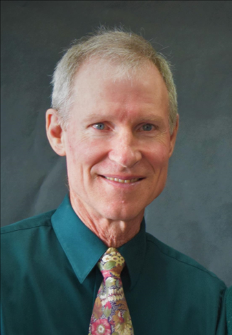 A middle-aged man with short blonde hair and blue eyes, smiling, wearing a teal shirt and a patterned tie, against a plain dark background.