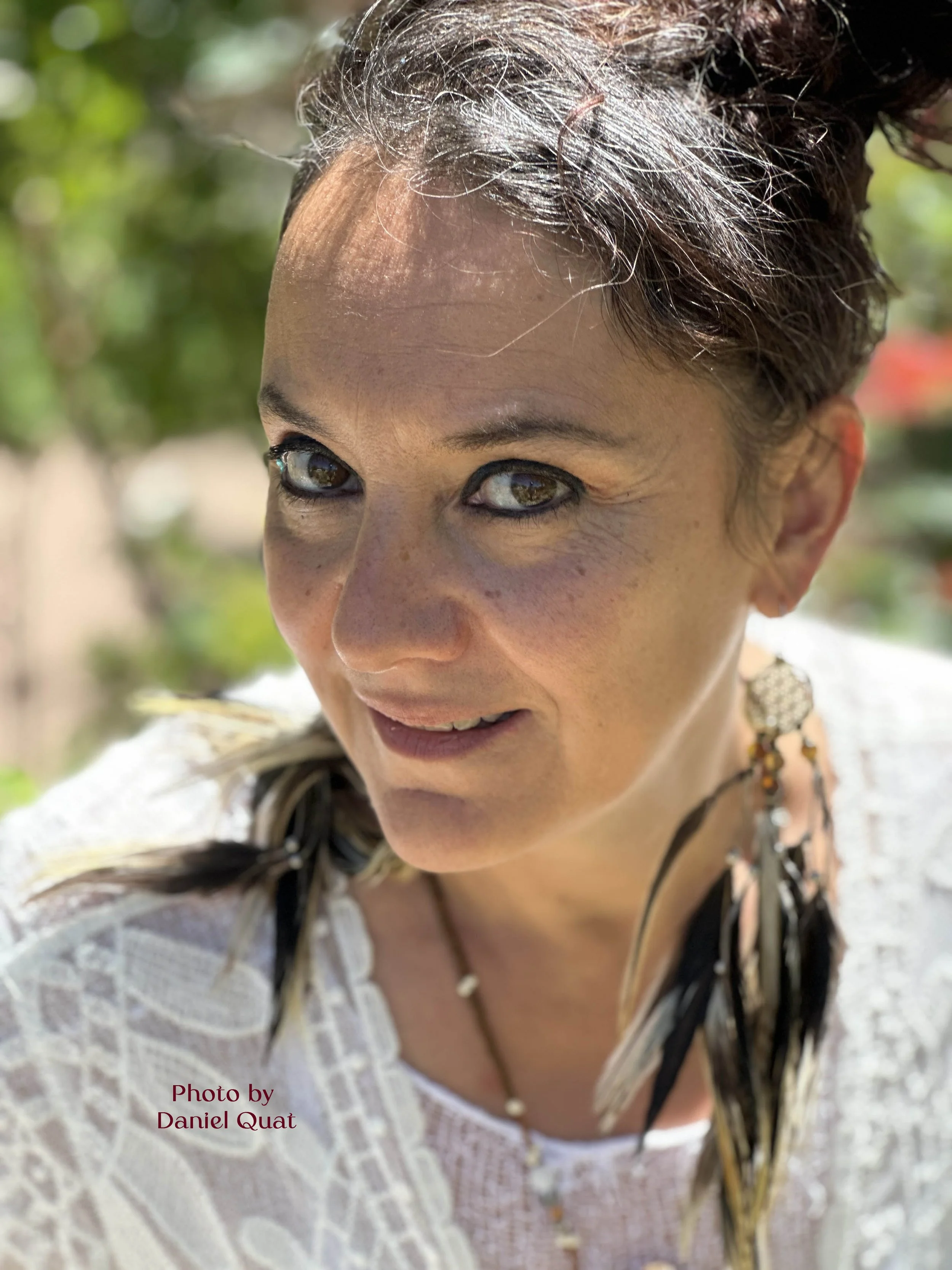Close-up of a woman with light skin, dark hair with feather details, and green eyes, outdoors with blurred green foliage in the background.