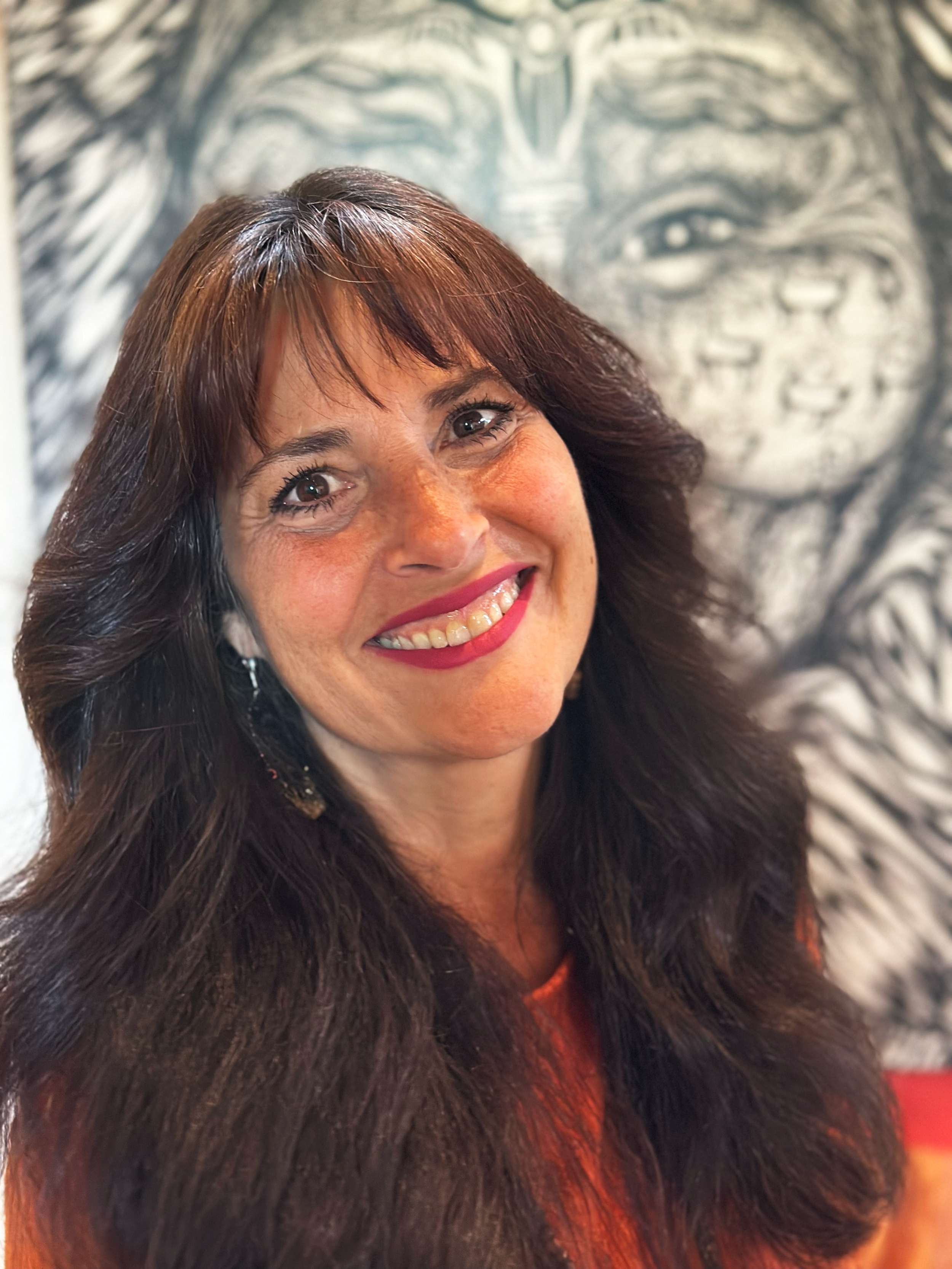 A woman, Rosy Aronson, with long dark brown hair and light skin smiling in front of a large black and white drawing of a Wisdom Keeper face.