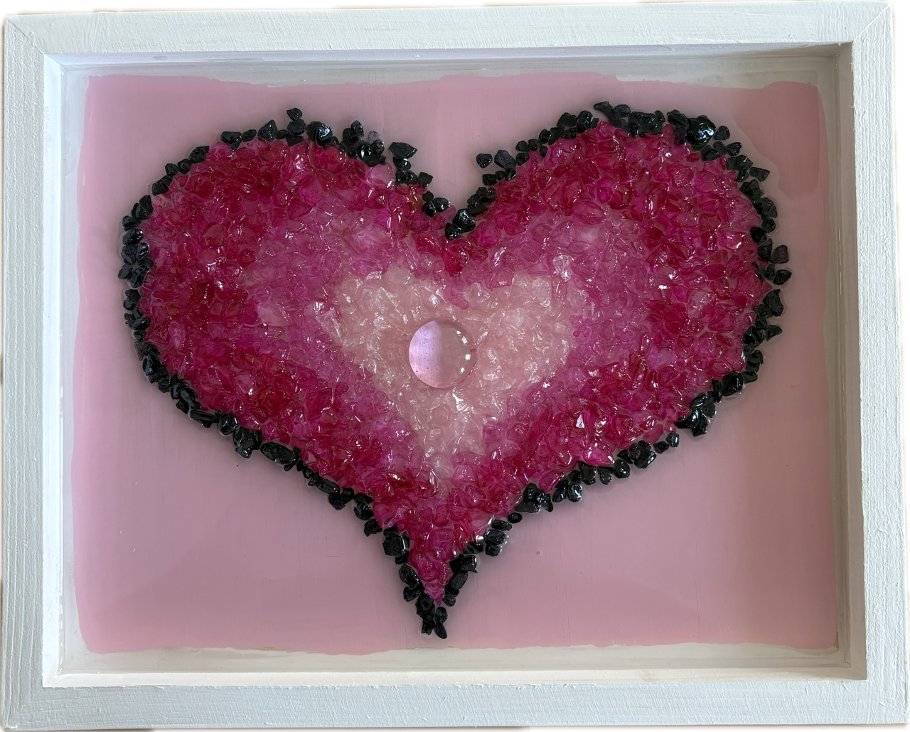 Decorative resin art using crushed glass heart shape made of pink, white, and black small stones inside a white frame with a pink background.