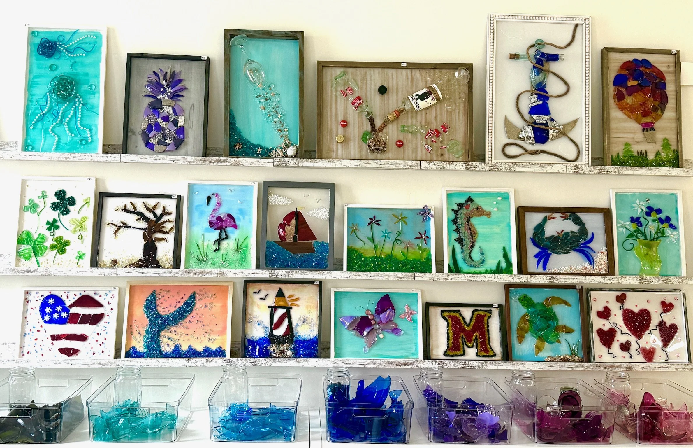 Display of various colorful glass resin art pieces mounted on shelves and framed, depicting marine life, abstract designs, and artistic representations of animals, with containers of colored glass at the bottom.