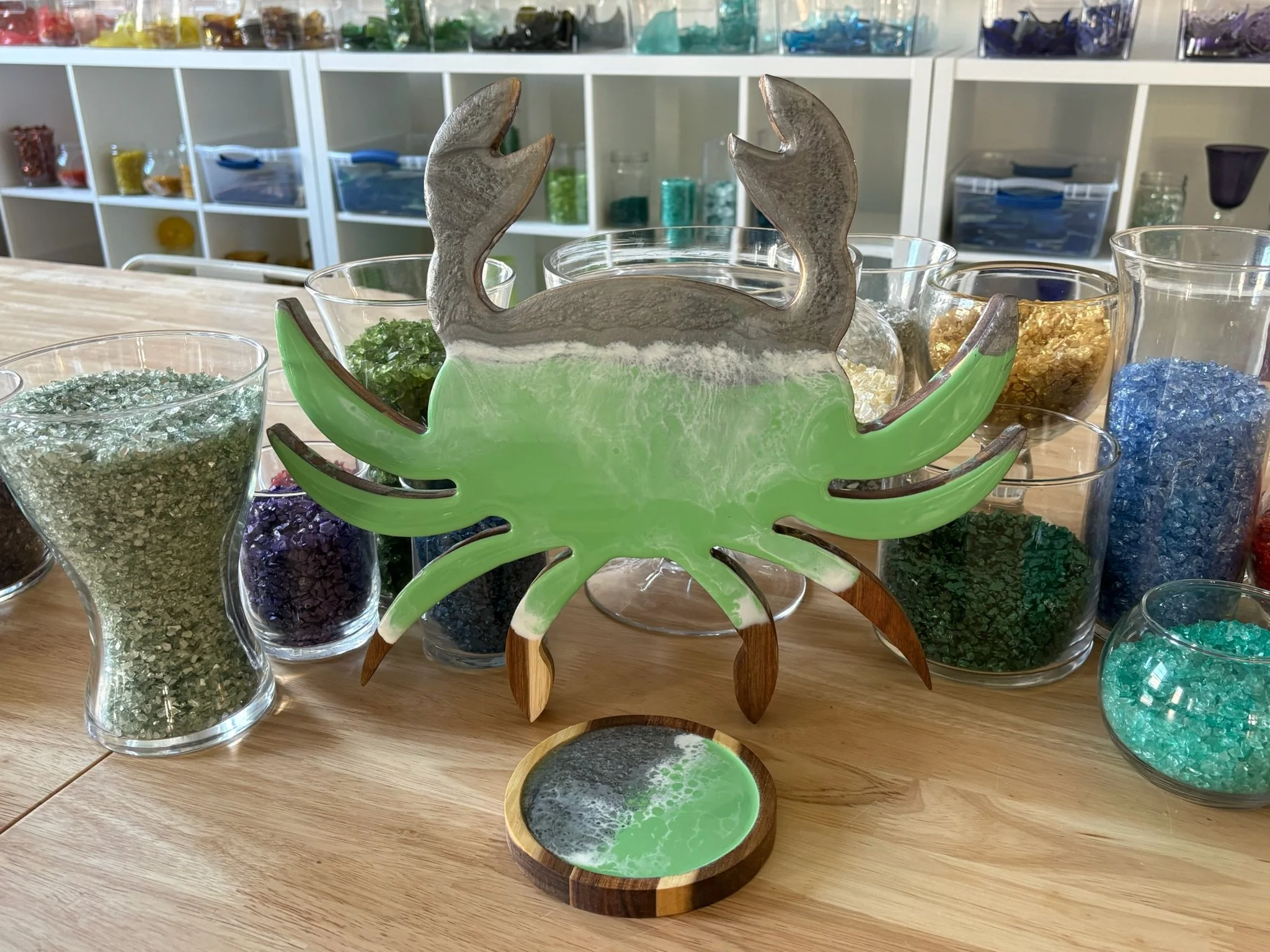 Colorful resin glass art piece of a crab with green and brown accents, surrounded by vases filled with various colored glass chips on a wooden table.