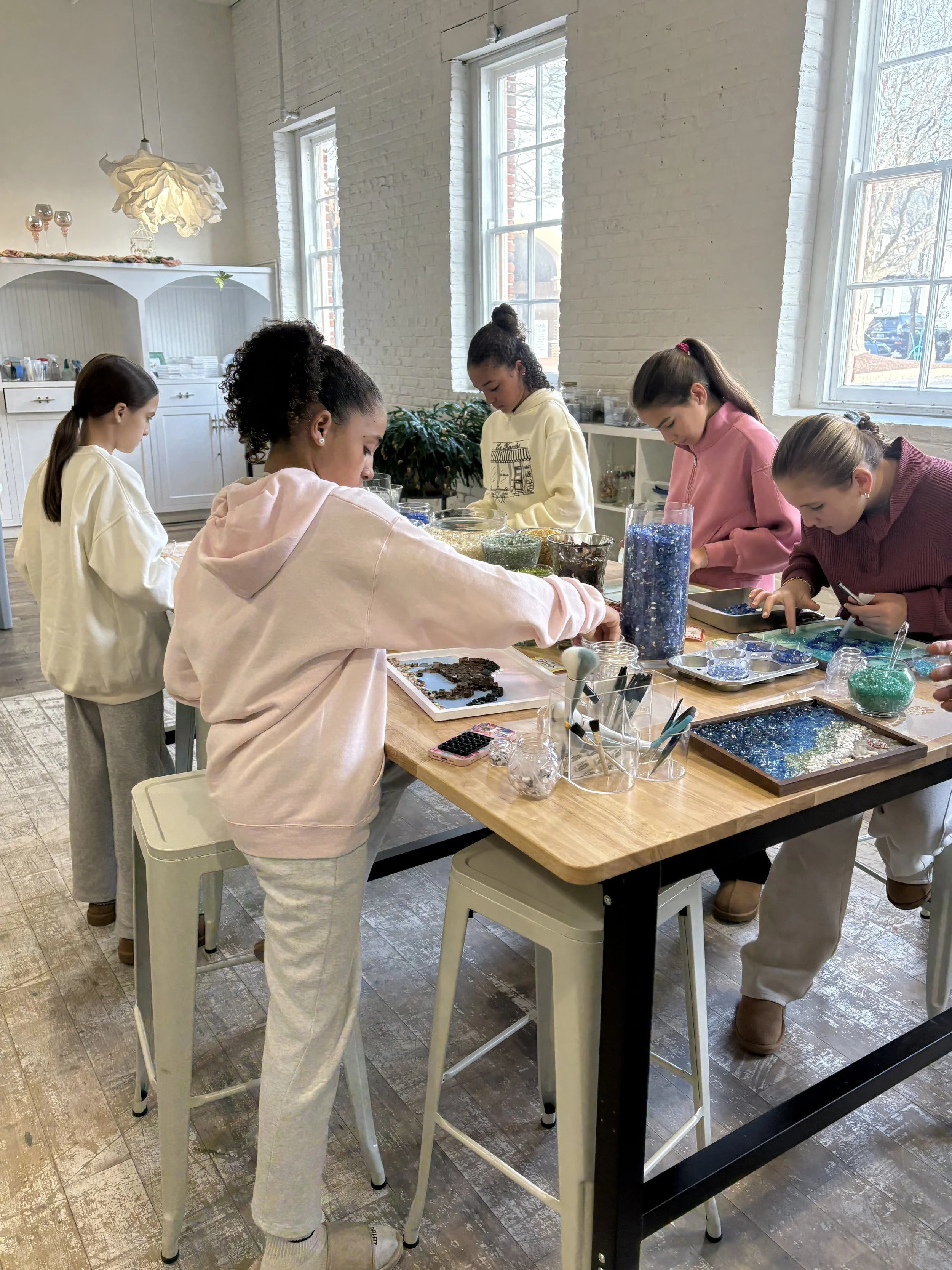 Private Birthday Party DIY workshop at The Resin Room in Annapolis MD. Five young girls gathered around a wooden table, working on arts and crafts projects using cut and crushed glass. DIY classes.