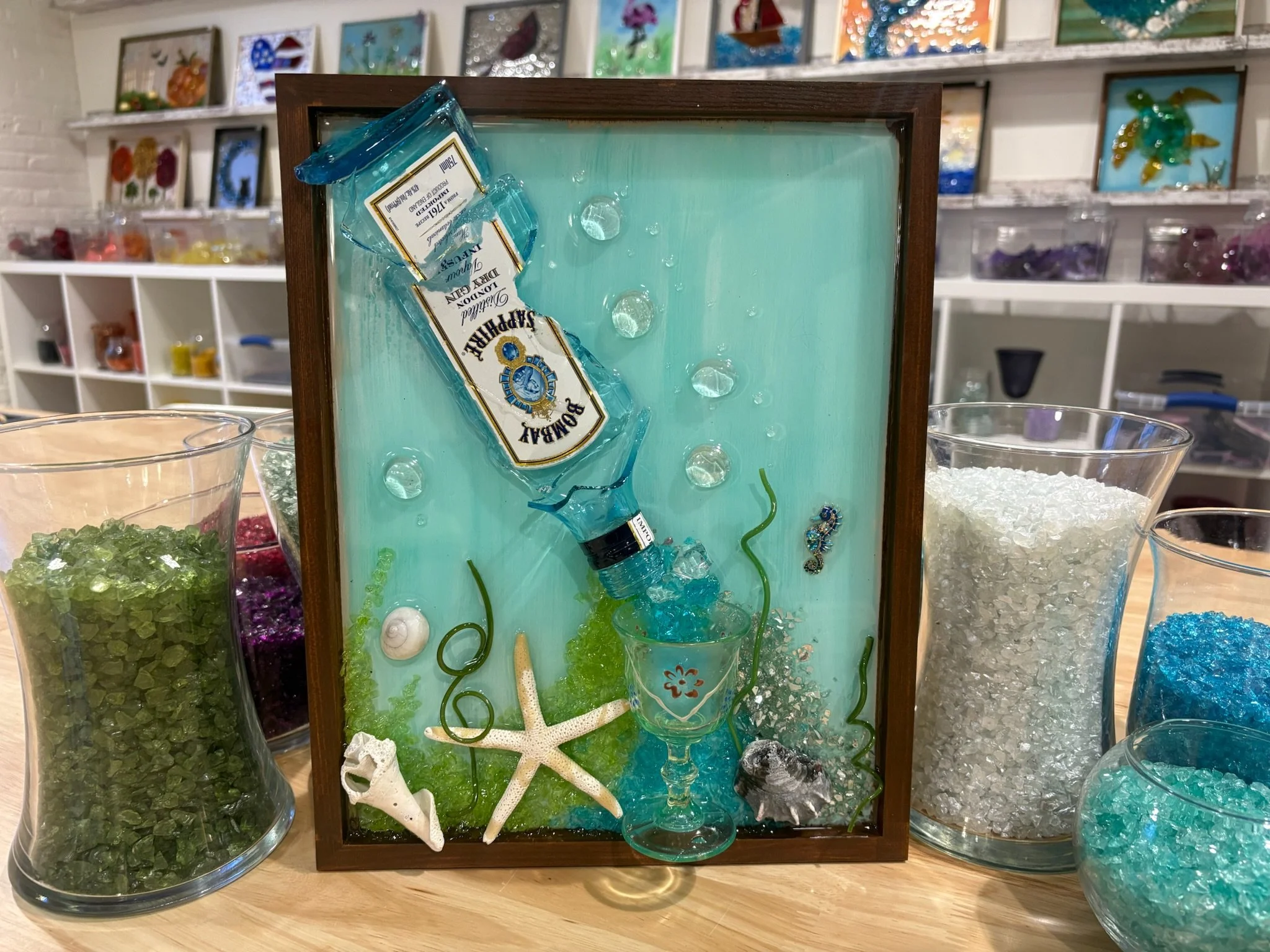 Resin art of shattered  'BOMBAY SAPPHIRE London Dry Gin' bottled and martini glass surrounded by decorative sea life items like a starfish, shells, and green coral.